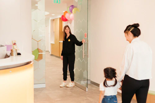 A woman opens a glass door, smiling toward another woman and a young girl holding hands and walking into a brightly lit, modern office with colorful balloons above the door.